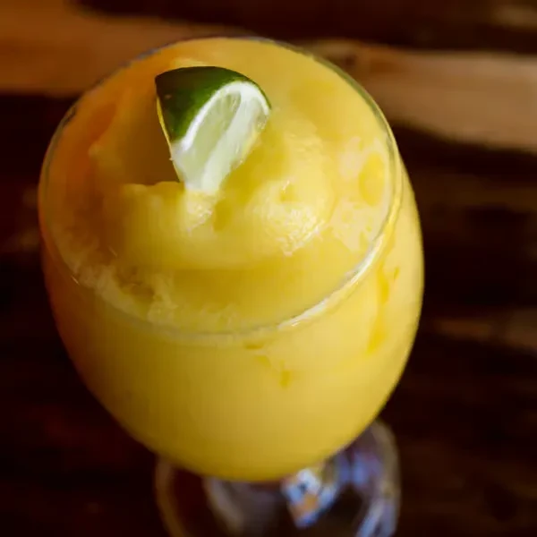 Yellow slushy drink with lime wedge in stemmed glass on dark background.