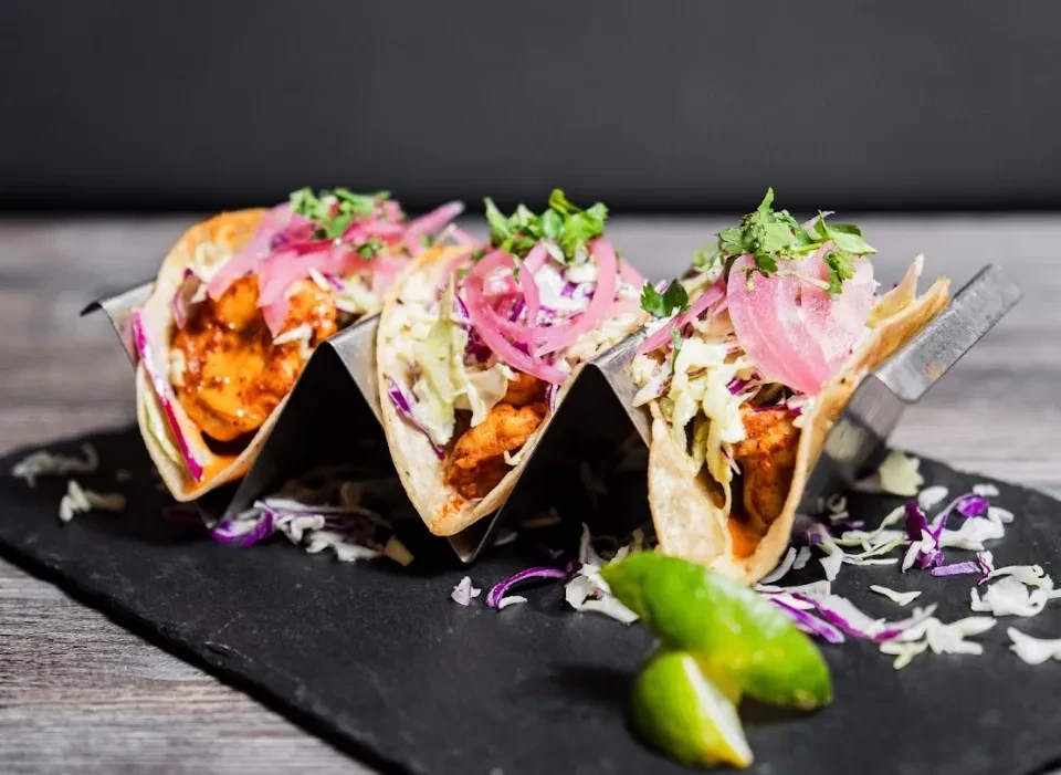 Three vibrant tacos in a metallic holder on dark slate, with lime.