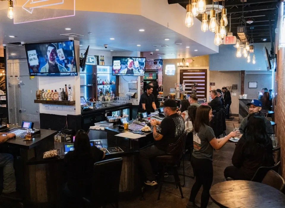 Busy modern bar with patrons, bartender, stocked shelves, and TVs.