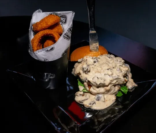 Piled burger with mushroom sauce, knife, and Tavern-branded onion rings.