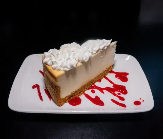 Cheesecake slice with whipped cream, red sauce, on a white plate.