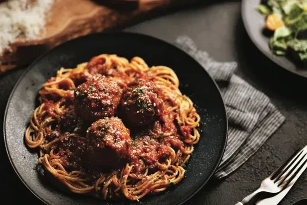 Close-up of spaghetti with meatballs, Parmesan, and herbs on a dark plate.