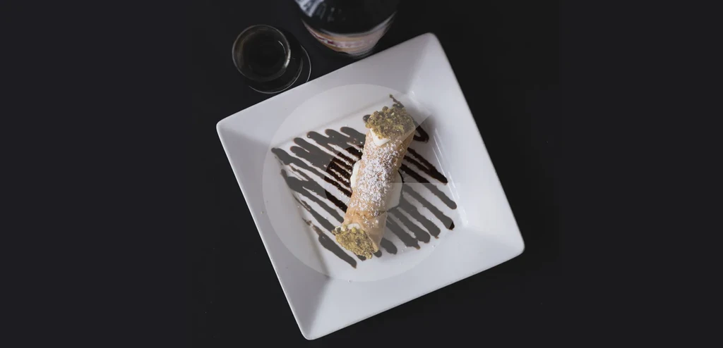 Cannoli with powdered sugar, pistachios, and chocolate drizzle on white plate.