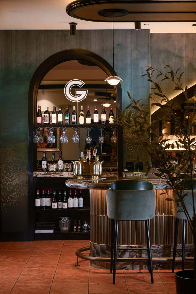 Chic bar with arched mirror, glowing 'G' reflection, wine, and velvet stools.