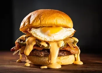 Close-up gourmet burger with melted cheese, bacon, jalapeños, and runny fried egg.