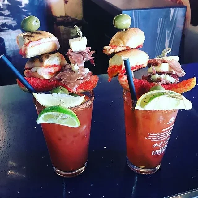 Two elaborately garnished cocktails with food skewers on a dark bar.