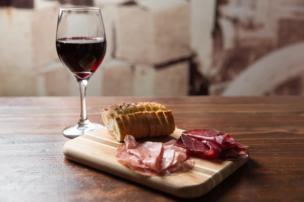 Close-up: red wine, sliced baguette, cured meats on a wooden board.