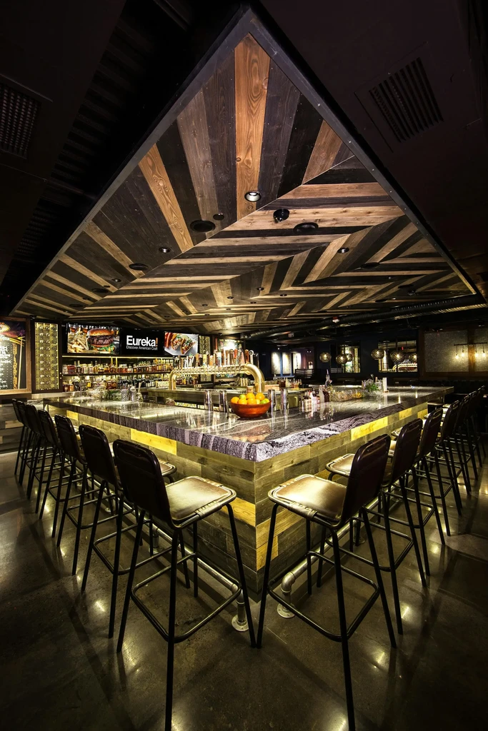 Modern bar with dark counter, patterned wood ceiling, and Eureka! TV.
