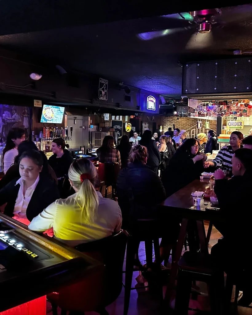 Bustling, dimly lit bar interior with patrons, a long bar, and colorful lights.