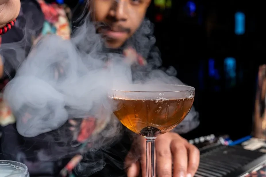 Dramatic smoke curls from an amber cocktail observed by a bartender.
