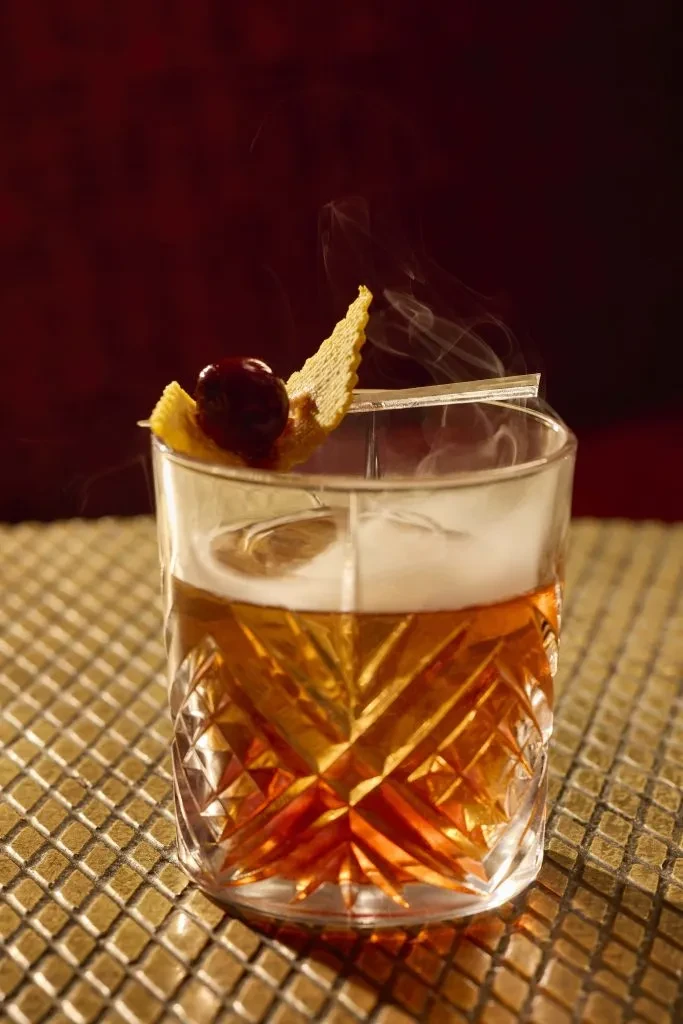 Ornate crystal cocktail with rising smoke, cherry, citrus, and wafer.