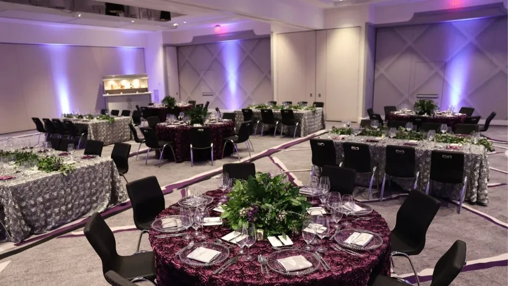 Elegantly set banquet hall with maroon, silver tablecloths, foliage, purple lighting.
