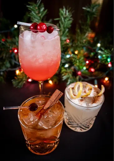 Three colorful, garnished cocktails, with blurred Christmas tree background.