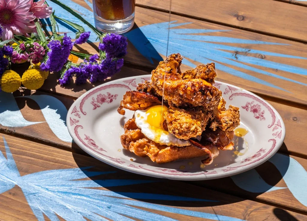 Plate of crispy chicken and waffles with egg, bacon, drizzled syrup.