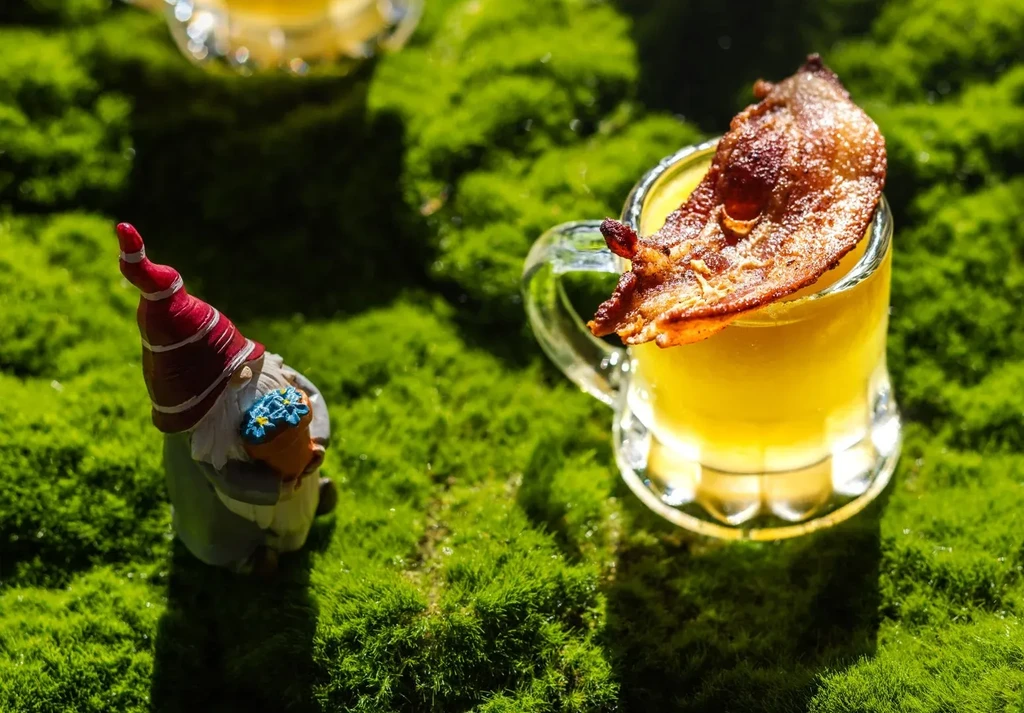 Yellow drink with bacon strip, next to a garden gnome on moss.