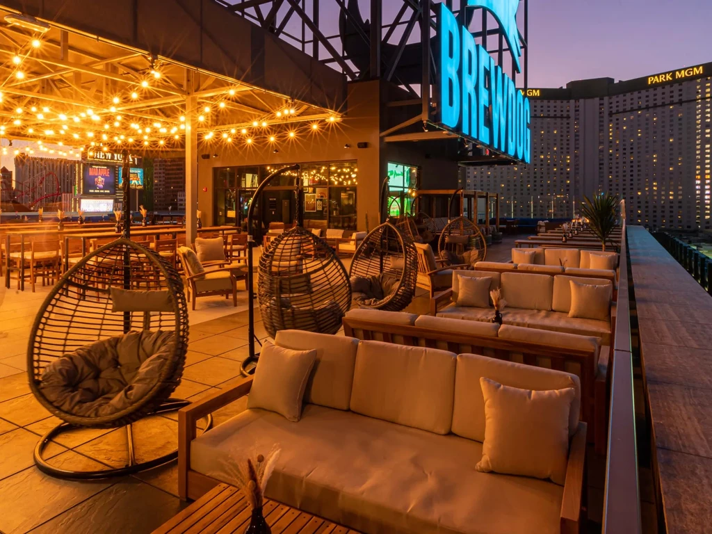 Rooftop bar with hanging chairs, string lights, 'BREW' sign, and city skyline.