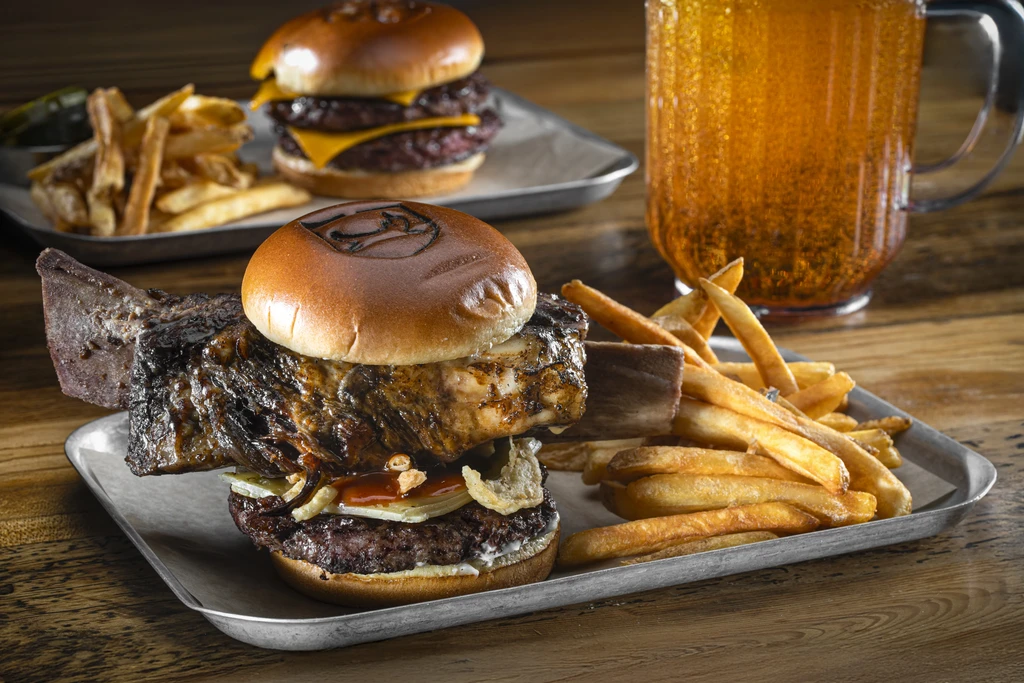 Large burger topped with bone-in meat, fries, and beer on wood.