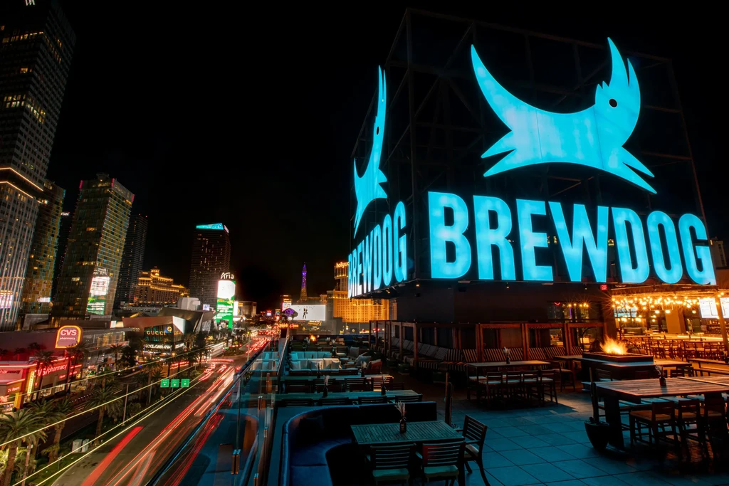 Glowing Brewdog sign above a lively rooftop bar, city night scene.