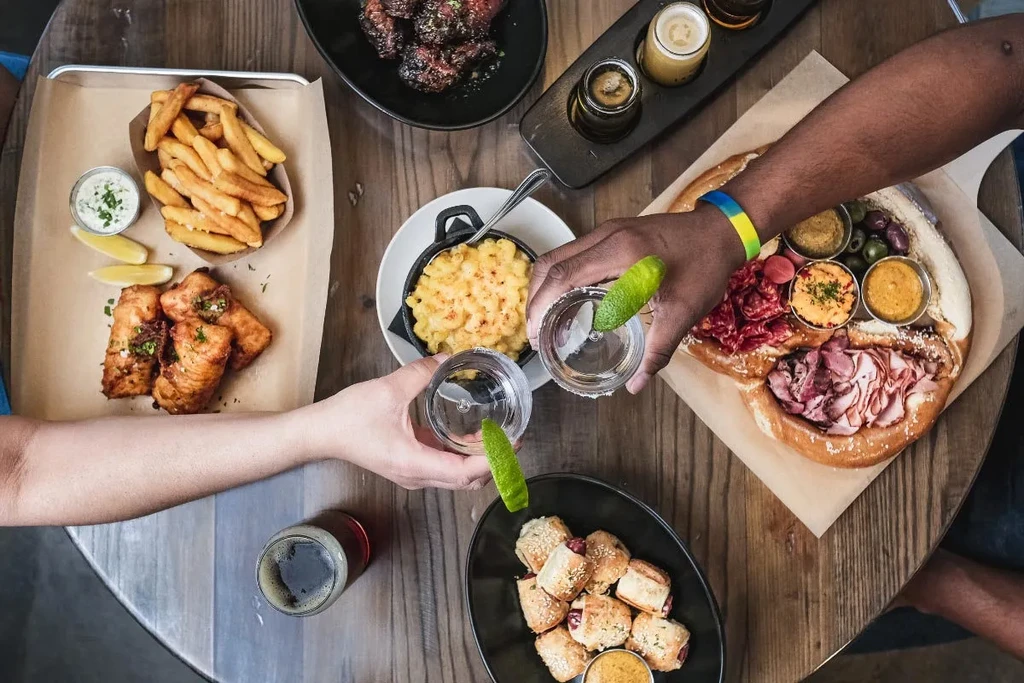 Overhead table with food, beer flight, and hands toasting drinks.