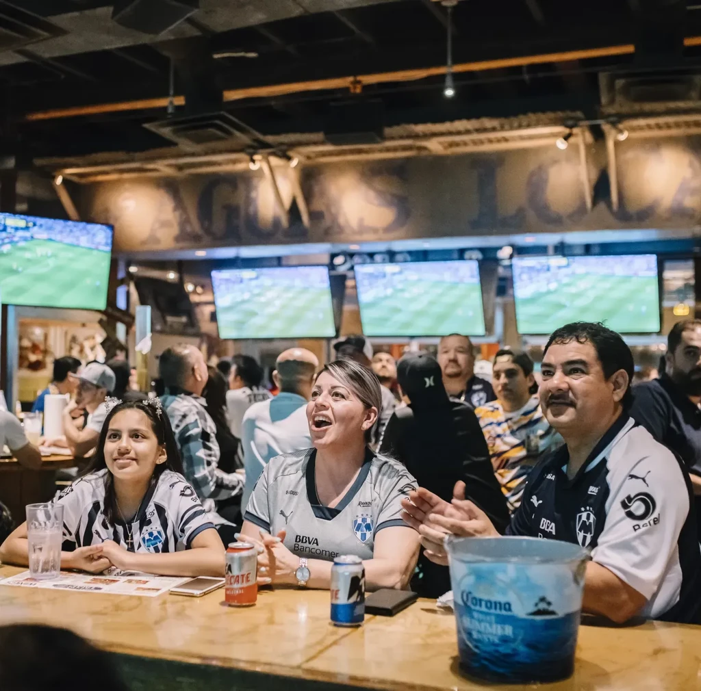 Three people in jerseys watch soccer intently on bar TVs.