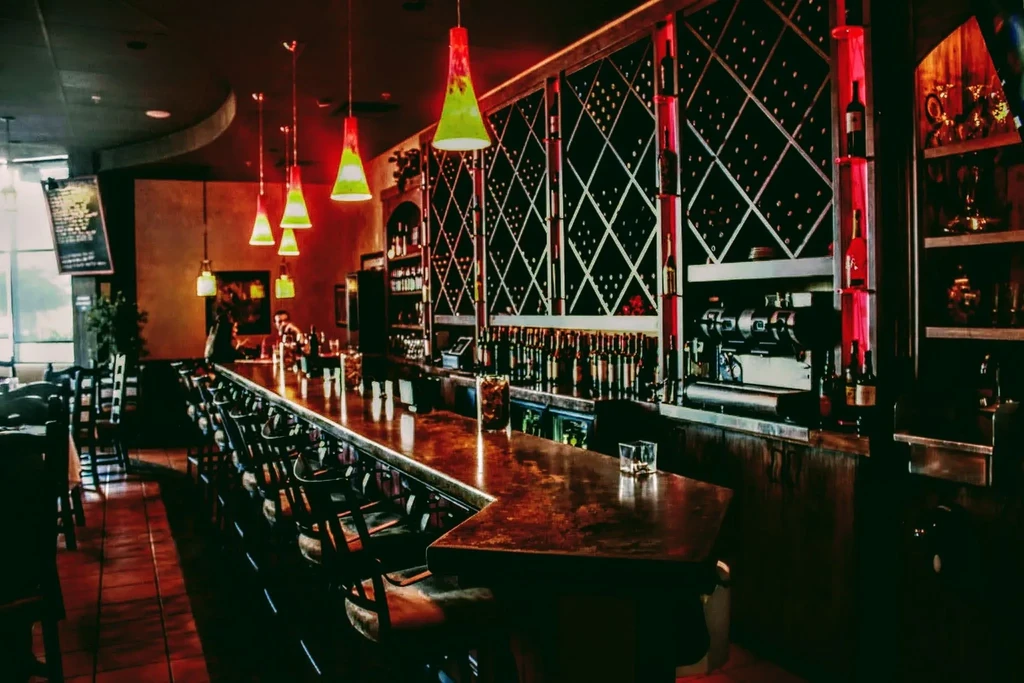 Upscale bar with long wooden counter, green pendant lights, red wine rack.