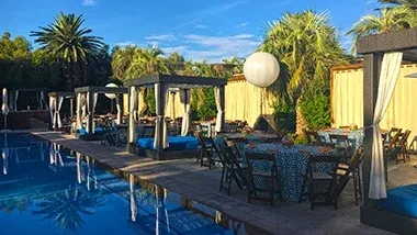 Outdoor resort pool with cabanas, patterned dining tables, and palm trees.