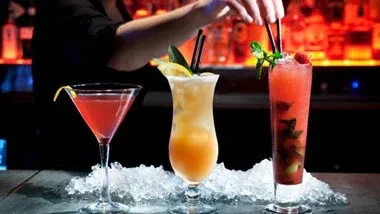 Three garnished cocktails on crushed ice, a bartender adjusts one.