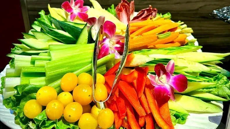 Platter with fresh raw vegetables, yellow fruits, orchid blossoms, and tongs.