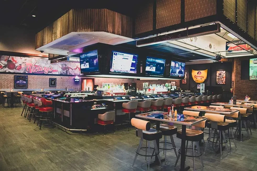 Modern sports bar interior with central bar, multiple TVs, and 'Victory's' mural.