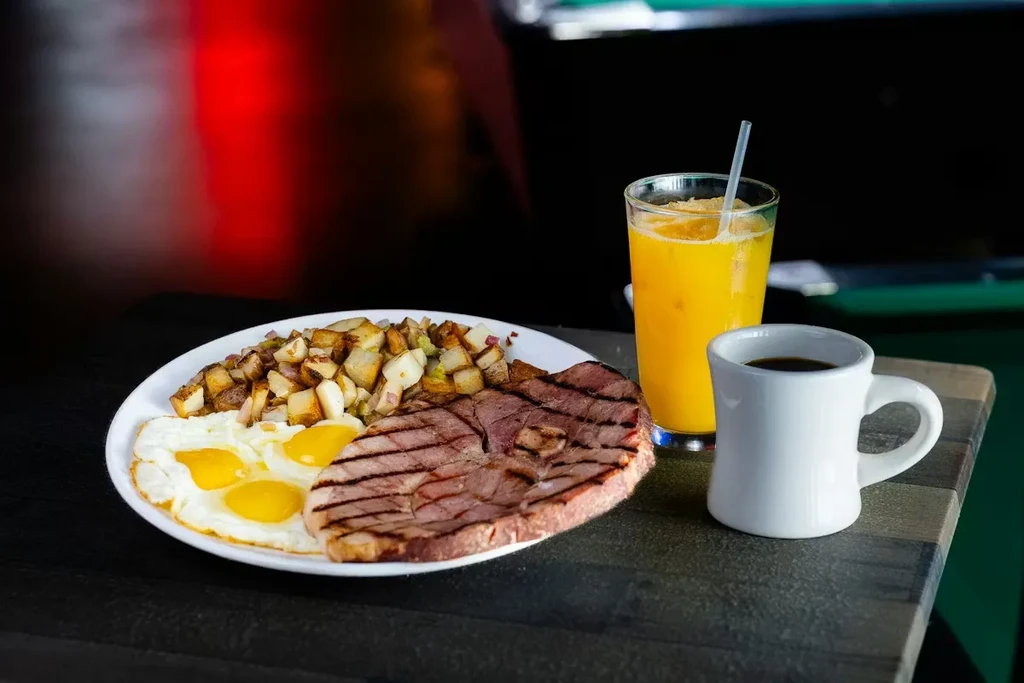 Hearty breakfast with fried eggs, grilled meat, home fries, juice, and coffee.