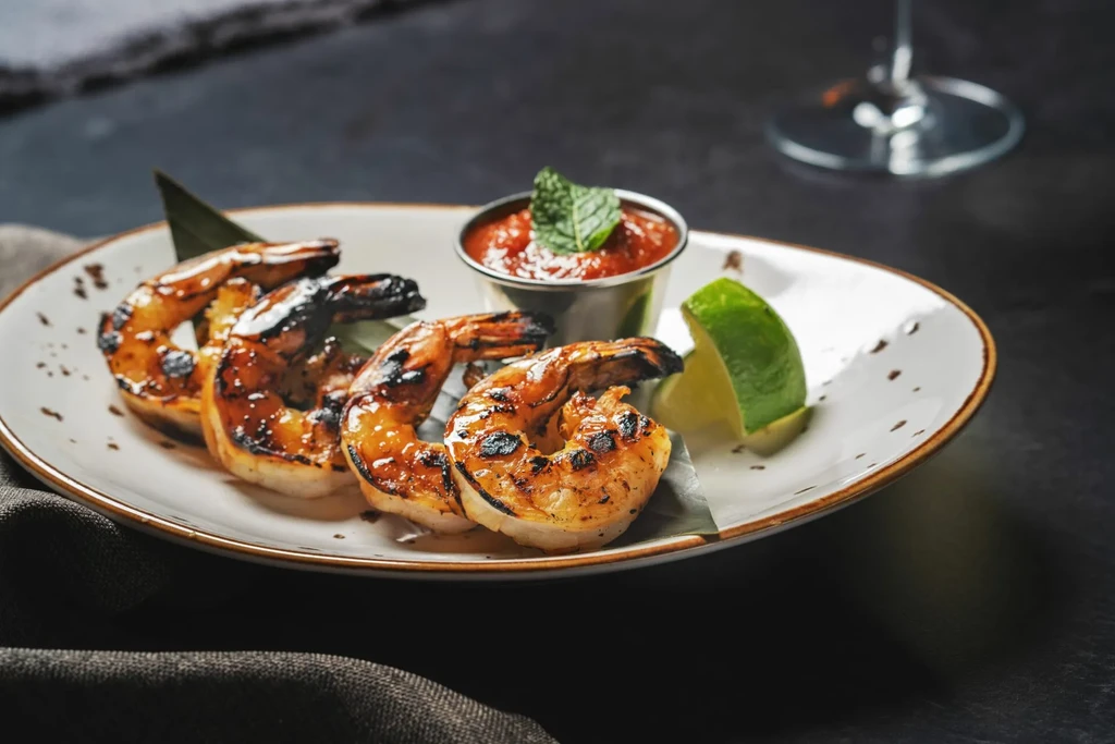 Four grilled shrimp with red sauce and lime on a rustic plate.