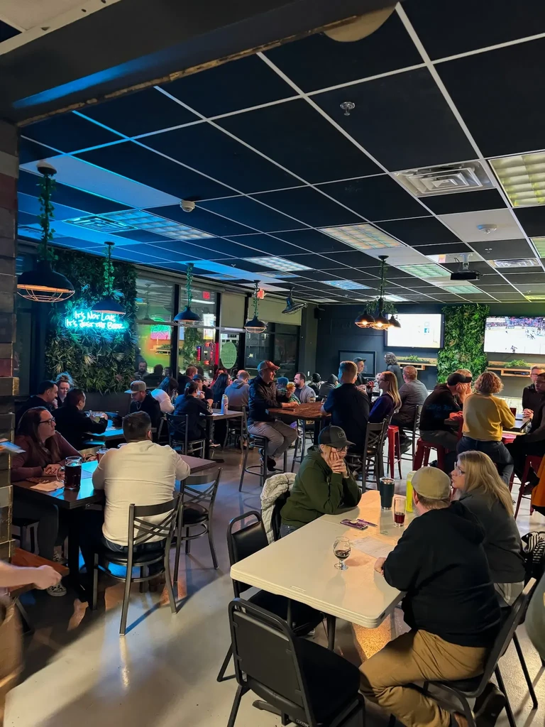 Bustling modern bar interior with patrons, a neon sign, and TVs.