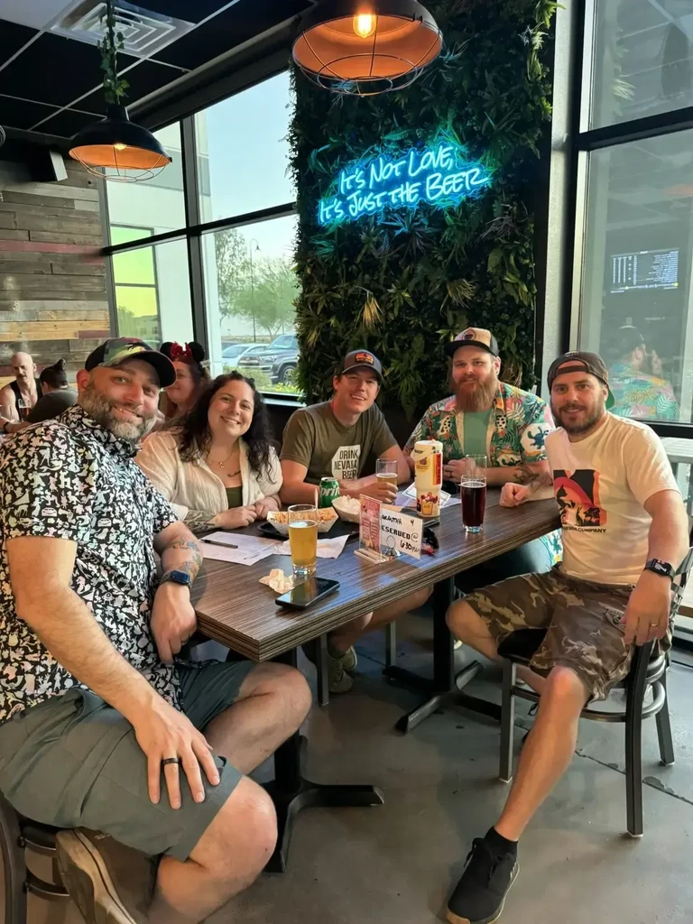 Five smiling people with drinks at a restaurant featuring a beer sign.