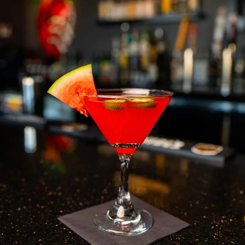 Red cocktail in martini glass with watermelon garnish on a bar.