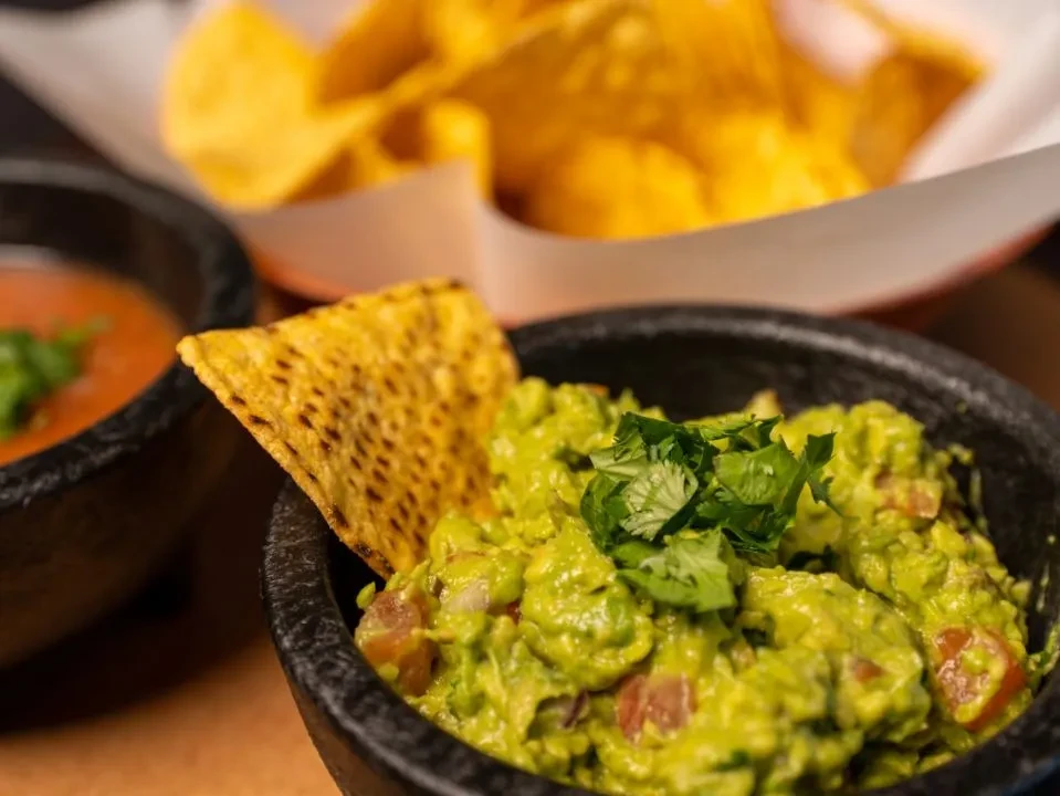 Close-up of chunky green guacamole with cilantro, a chip, salsa nearby.