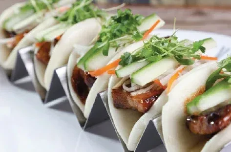 Open-faced pork belly bao buns in silver stands with pickled vegetables.