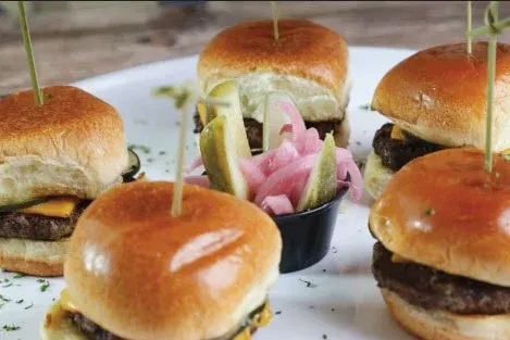 Five mini cheeseburger sliders with pickle spears and pink pickled onions.
