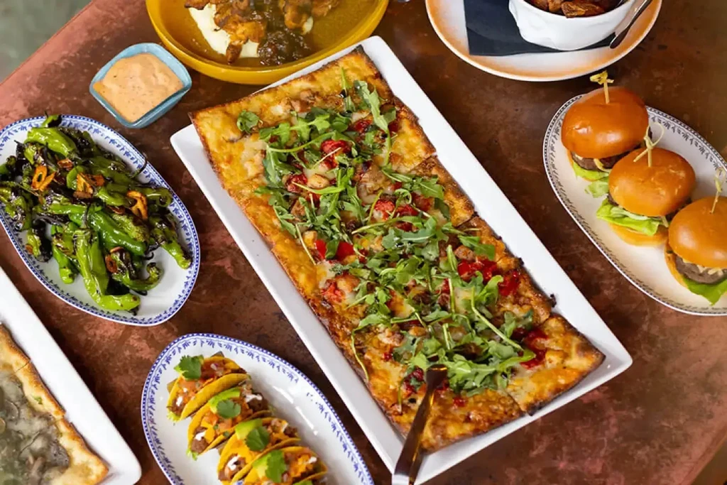 Flatbread, tacos, sliders, and shishito peppers on a reddish-brown table.