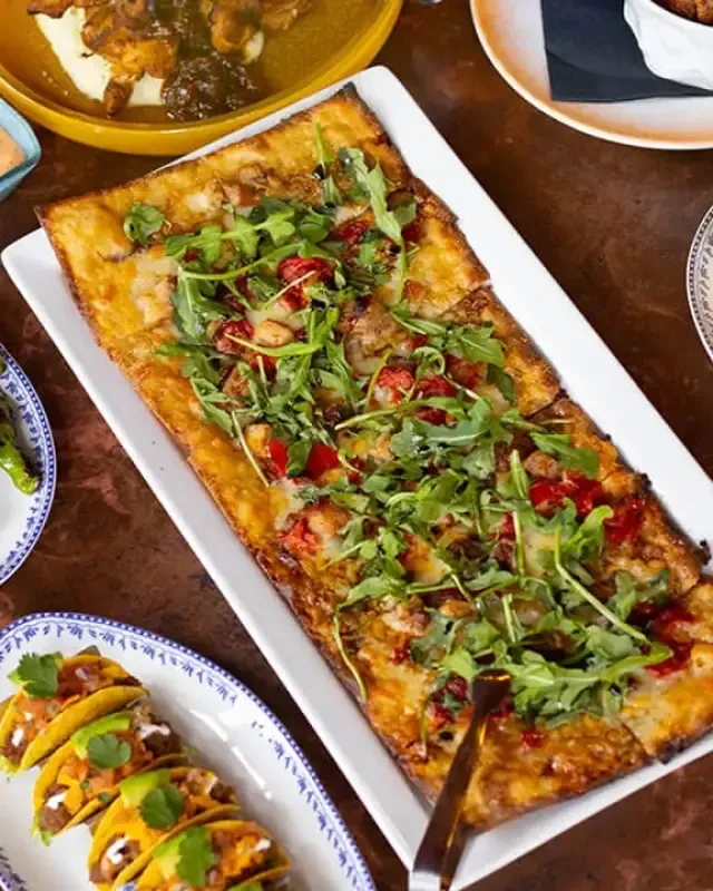 Overhead shot of a flatbread pizza, mini tacos, and two additional dishes.