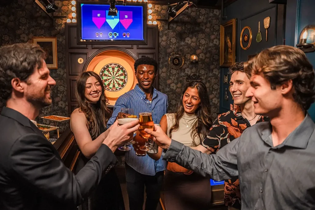 Six smiling young adults toast drinks in a bar with a dartboard.