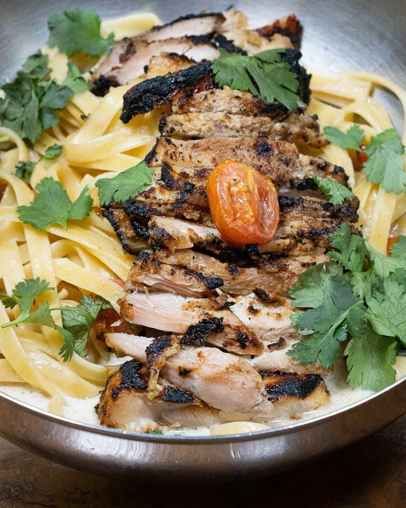 Grilled chicken and creamy pasta garnished with cilantro and tomato.