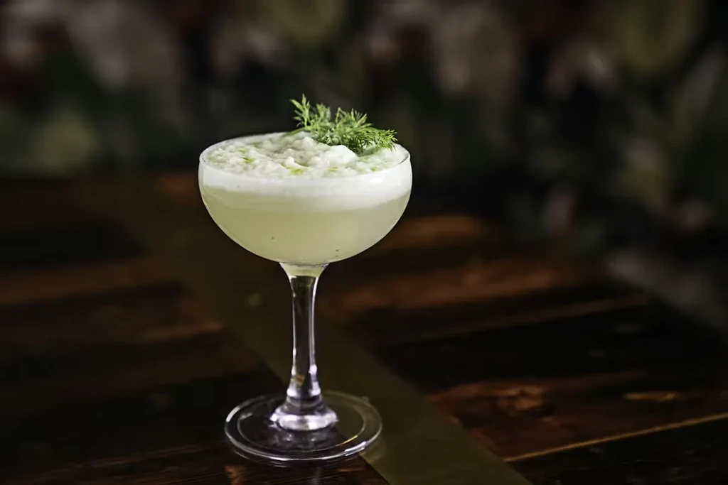 Pale green cocktail in coupe glass with frothy top and herb garnish.