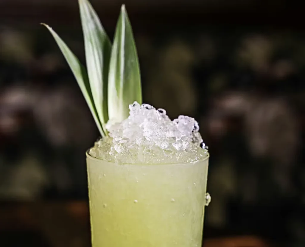 Tall glass of greenish-yellow drink with crushed ice and green leaf garnish.
