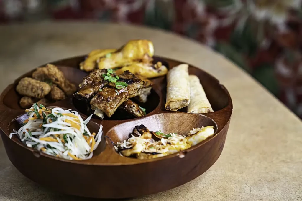 Wooden platter featuring grilled ribs, spring rolls, fried food, and slaw.