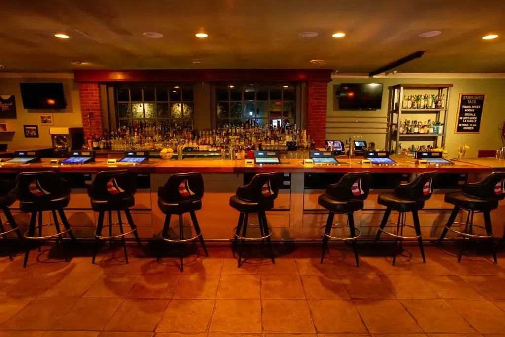 Empty bar counter with liquor, digital screens, logo stools, oyster menu.