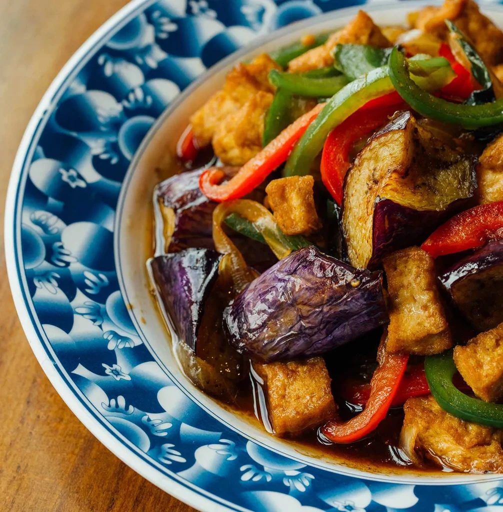 Stir-fried eggplant, tofu, bell peppers in dark sauce, white bowl.
