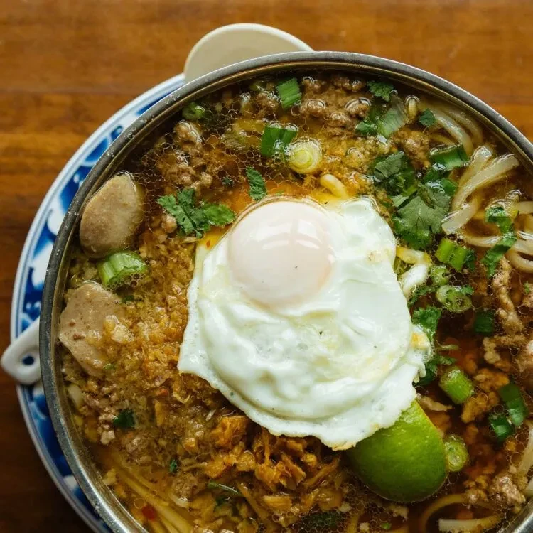 Overhead noodle soup with fried egg, meat, lime, and herbs.