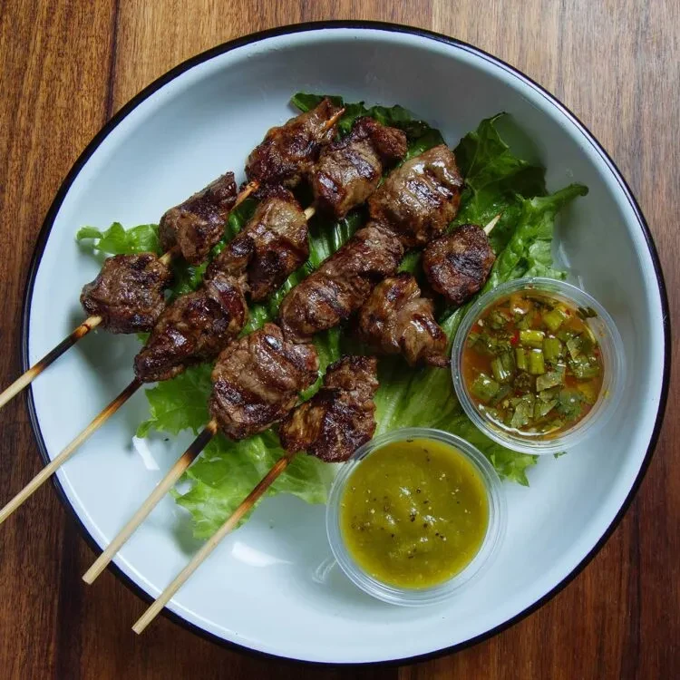 Grilled meat skewers, lettuce, and two sauces in a bowl on wood.