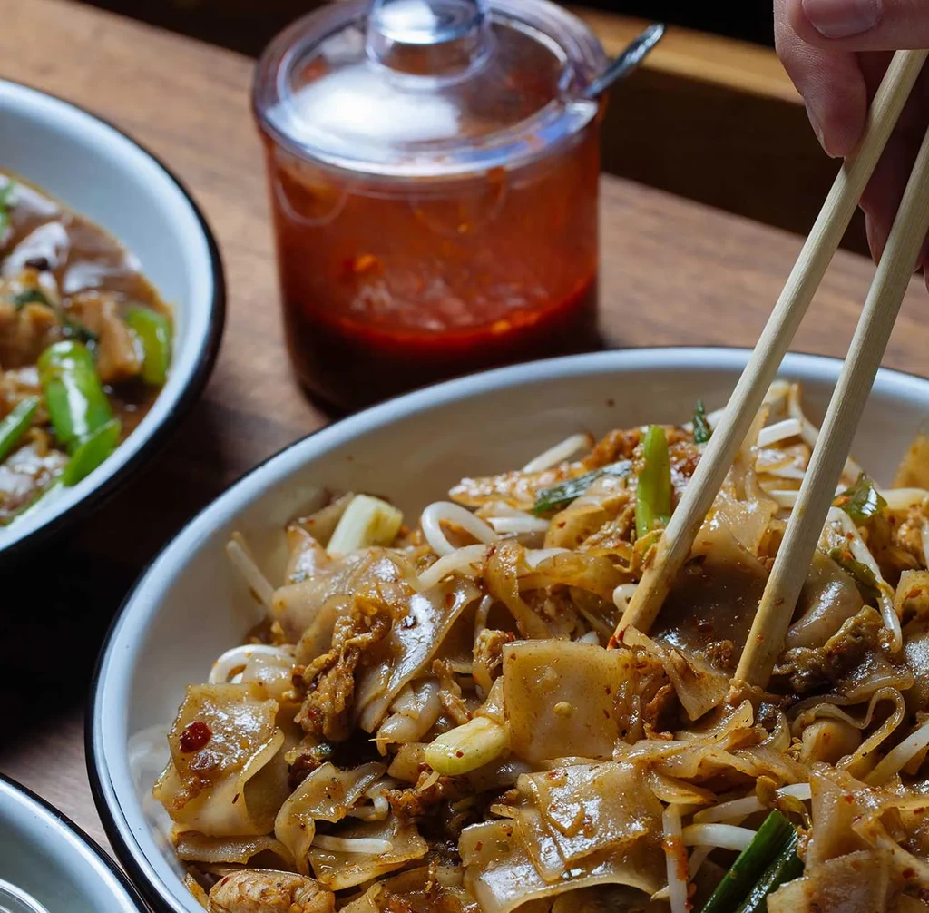 Chopsticks lift broad stir-fried noodles with egg from a white bowl.
