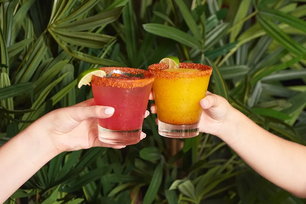 Two hands hold red and yellow drinks with spiced rims and lime.